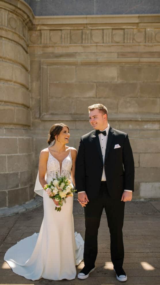 A bride in a white dress holding a bouquet and a groom in a black tuxedo stand hand in hand, smiling at each other, against a stone building background. by Justin Salem Meyer