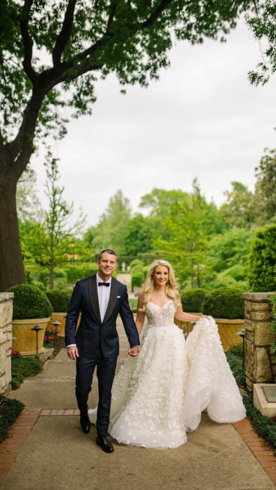 A bride in a long white gown and a groom in a dark suit walk hand in hand down a garden pathway, surrounded by lush greenery and stone walls, under a large tree on a cloudy day. by Justin Salem Meyer