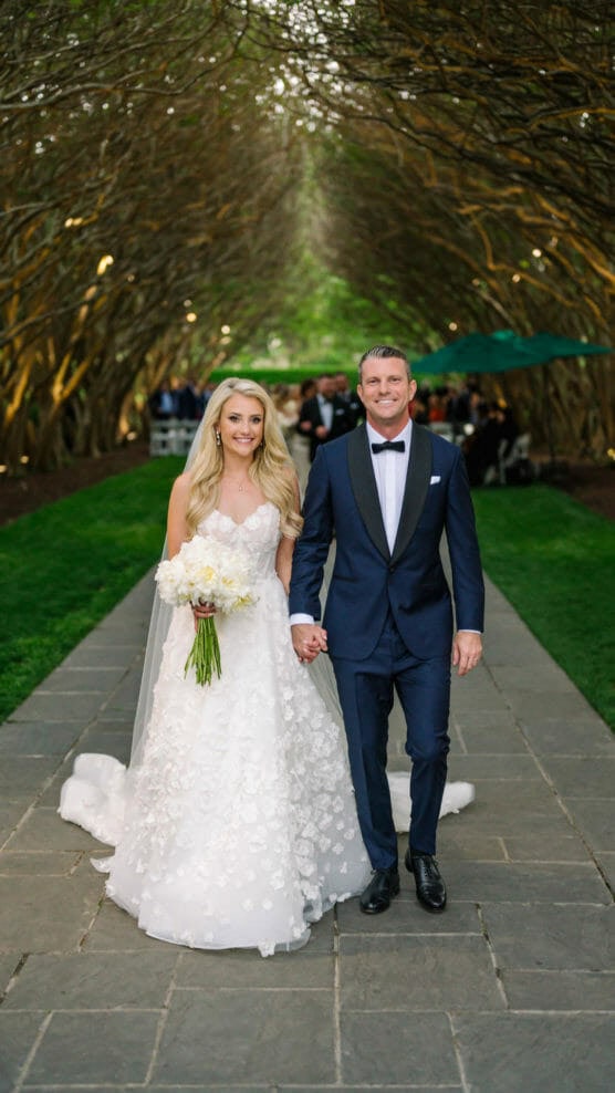 A bride in a white gown holding a bouquet and a groom in a black tuxedo walk hand in hand down a stone path under an archway of trees at an outdoor wedding. by Justin Salem Meyer