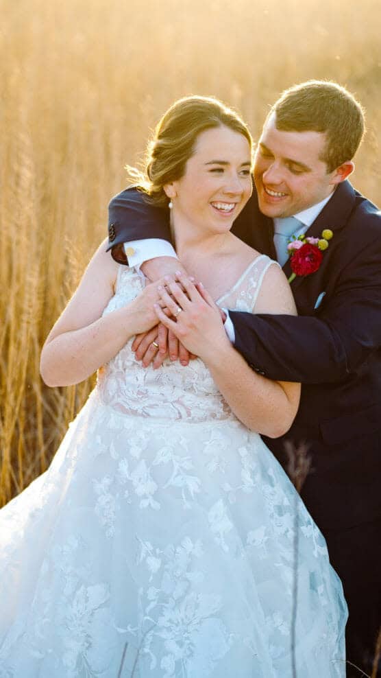 A bride in a white lace gown and a groom in a dark suit embrace and smile at each other in a sunlit field of tall golden grass. by Justin Salem Meyer
