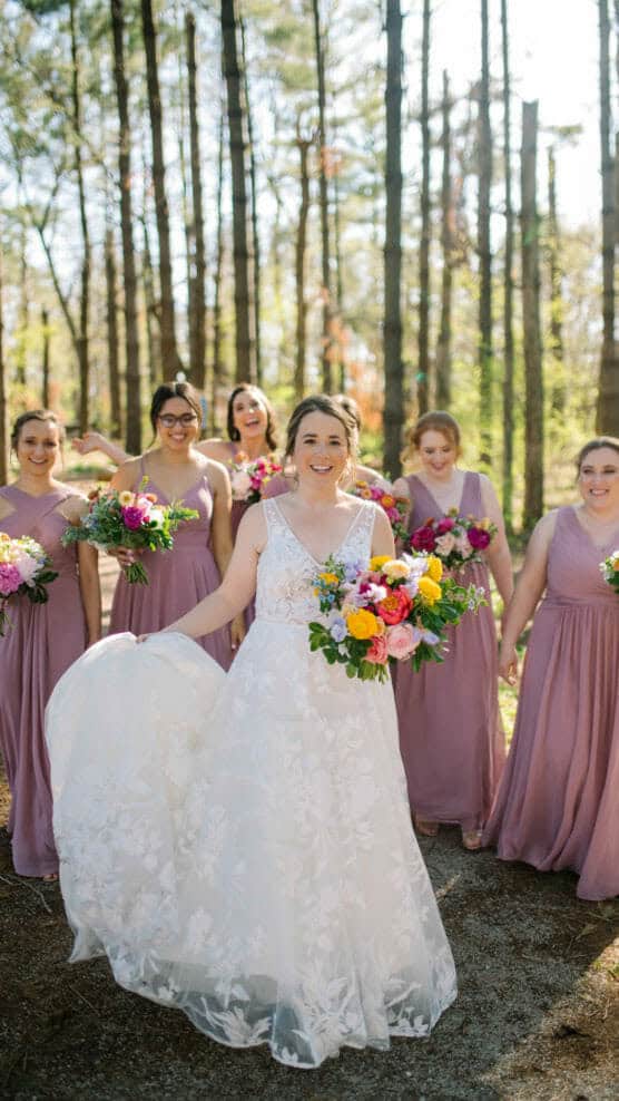 A bride in a white, floral lace gown holds a colorful bouquet and smiles, surrounded by five bridesmaids in matching mauve dresses, all holding bouquets, standing outdoors among tall trees. by Justin Salem Meyer