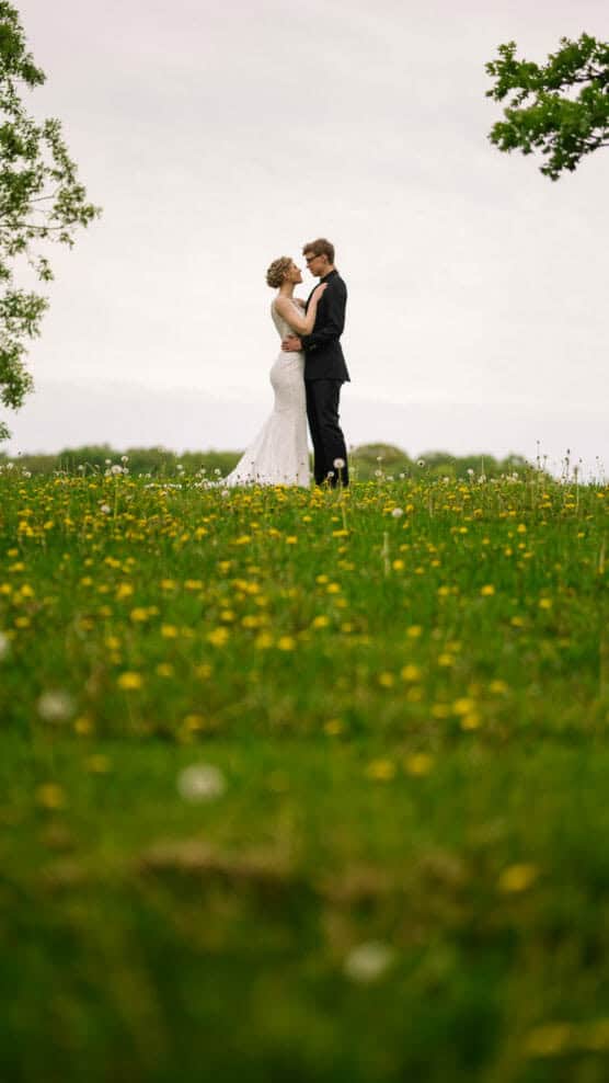 A couple dressed in wedding attire stands close together on a grassy hill filled with yellow wildflowers, with green trees and a cloudy sky in the background. by Justin Salem Meyer