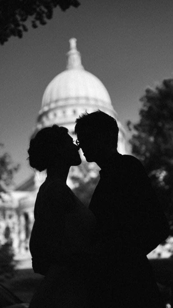 Silhouette of a couple about to kiss, standing close together with a large domed building in the background, framed by trees. by Justin Salem Meyer