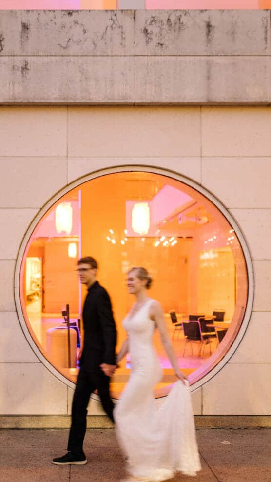 A couple holding hands walks past a large circular window glowing with warm orange light; the woman wears a white dress, and both appear slightly blurred in motion. by Justin Salem Meyer