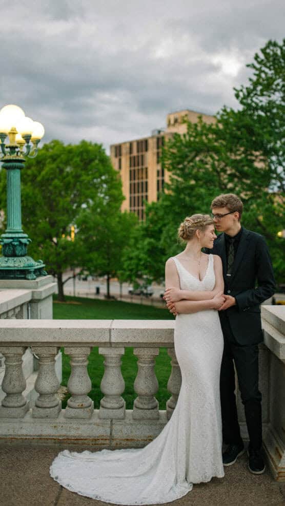 A bride in a white dress and a groom in a black suit stand close together on a stone balcony, surrounded by greenery and an ornate lamp post, with a city building in the background. by Justin Salem Meyer