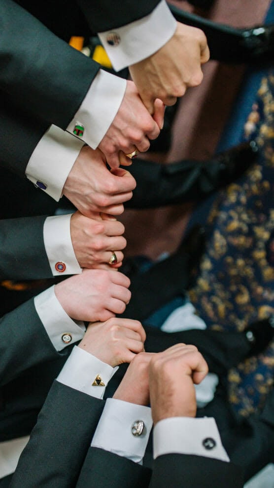 Six people in dark suits and white dress shirts stand in a circle, stacking their fists together in the center. Their cufflinks are visible, each with a different design. The gesture suggests unity or teamwork. by Justin Salem Meyer