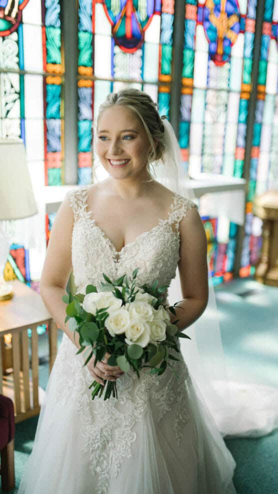 A smiling bride in a lace wedding dress holds a bouquet of white roses and greenery, standing in front of colorful stained glass windows in a sunlit room. by Justin Salem Meyer