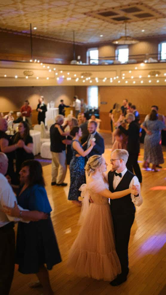 A couple in formal attire dances together in the center of a warmly lit ballroom, surrounded by other couples dancing. The room has string lights and wooden floors, creating a festive and joyful atmosphere. by Justin Salem Meyer