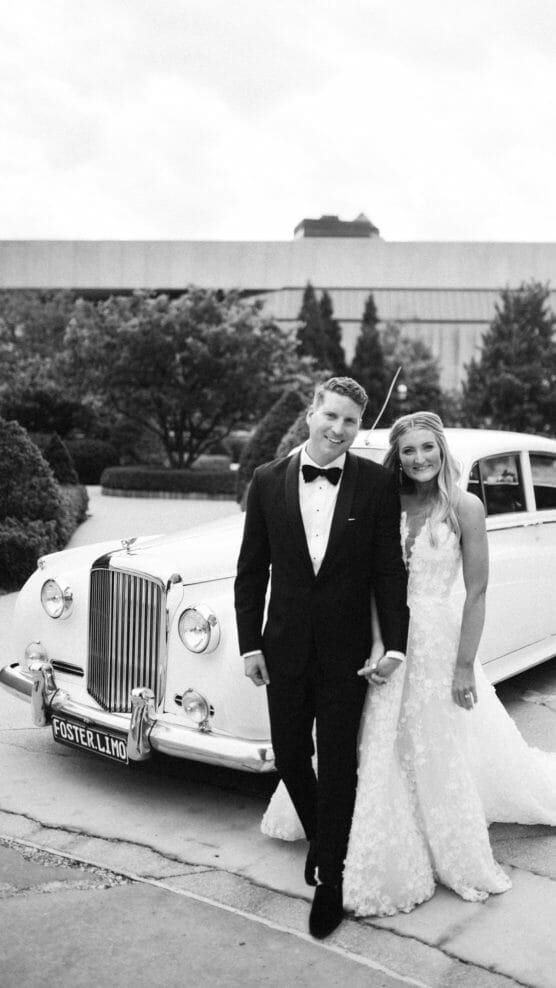 A smiling bride and groom in formal wedding attire stand together in front of a classic white car with a FOSTER LIMO license plate. Trees and a building are visible in the background. by Justin Salem Meyer