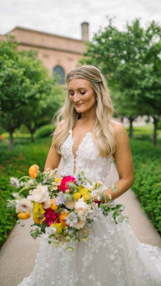 A bride in a white lace gown stands on a garden path holding a vibrant bouquet of colorful flowers, with green trees and a brick building in the background. She looks down and smiles softly. by Justin Salem Meyer