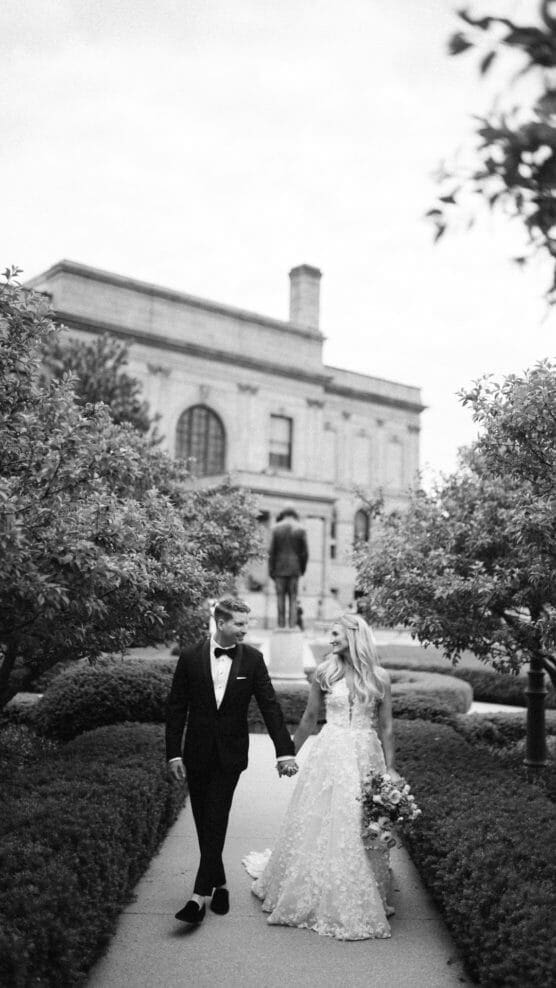 A bride in a white gown and a groom in a black suit hold hands while walking on a garden path, surrounded by hedges, with a stately building in the background. by Justin Salem Meyer