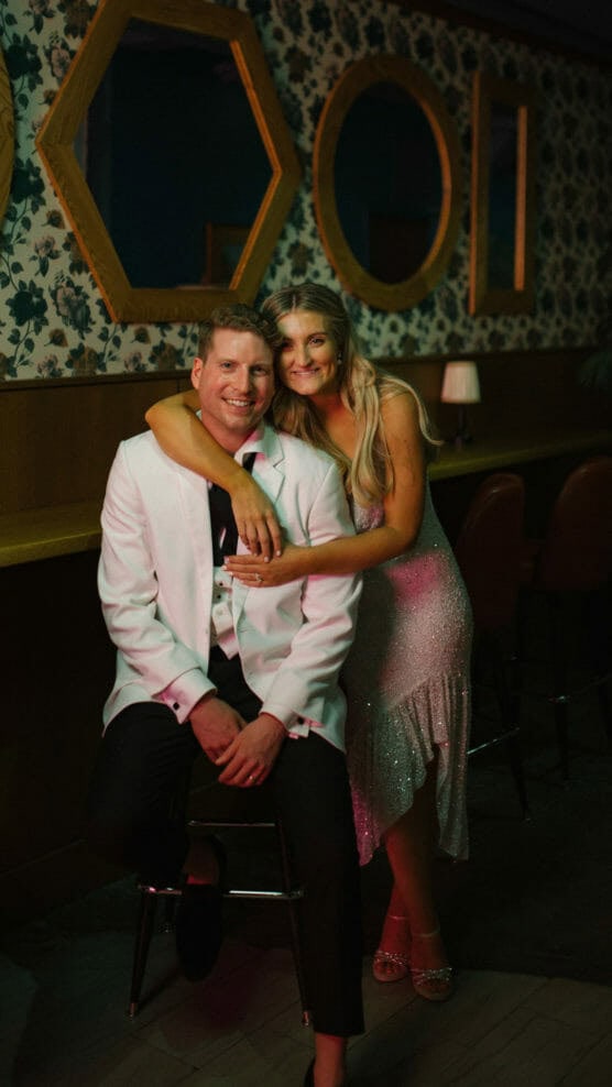 A smiling couple poses together indoors; the man sits on a barstool wearing a white tuxedo jacket, and the woman stands beside him in a sparkly dress with her arm around his shoulders. Geometric mirrors and floral wallpaper are behind them. by Justin Salem Meyer