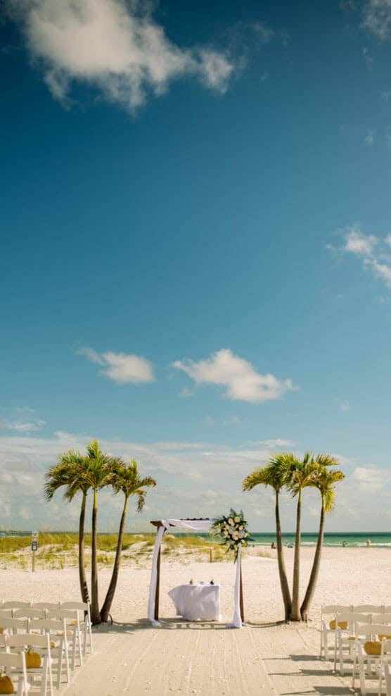 A wedding altar decorated with flowers stands on a sandy beach between two clusters of palm trees under a blue sky with scattered clouds. Rows of white chairs line the aisle in the foreground. by Justin Salem Meyer