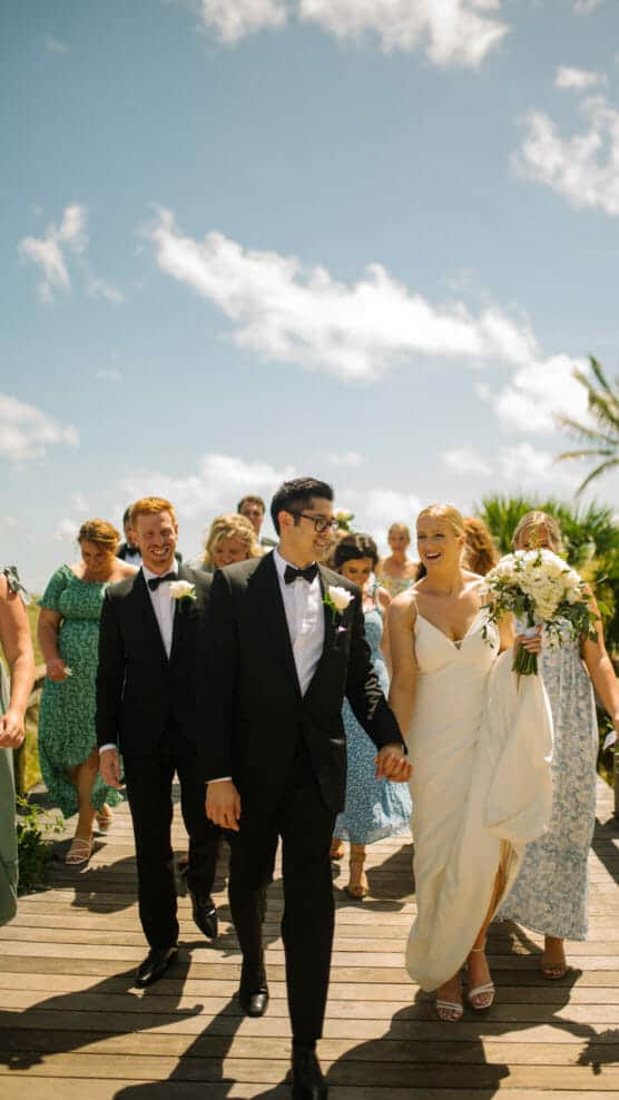 A bride in a white dress and groom in a black tuxedo walk hand in hand outside, smiling, surrounded by bridesmaids in blue dresses and groomsmen in suits, under a sunny, blue sky. by Justin Salem Meyer