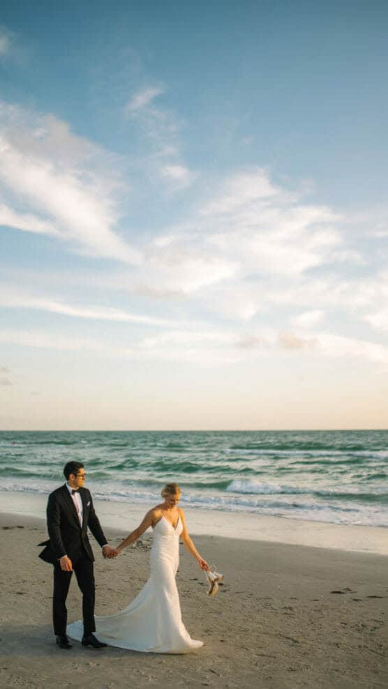 A bride in a white dress holds her shoes and walks barefoot on a sandy beach, holding hands with a groom in a black suit. The ocean waves and blue sky with clouds are in the background. by Justin Salem Meyer