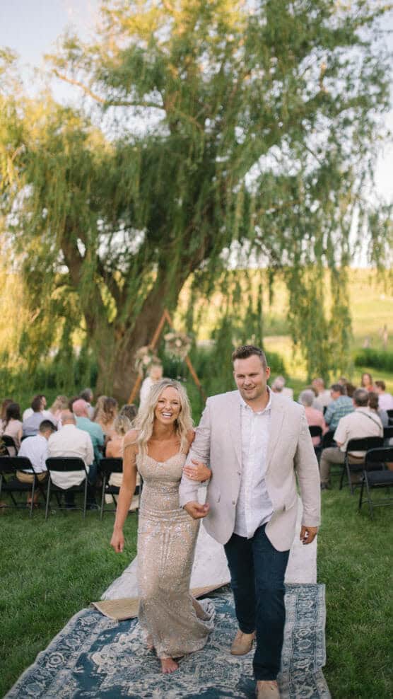 A smiling couple walks arm-in-arm down an outdoor aisle lined with rugs, with guests seated behind them under a large tree on a sunny day. The woman wears a sparkly dress; the man has a light jacket and dark pants. by Justin Salem Meyer