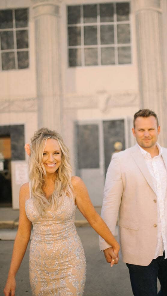 A smiling woman in a sparkly beige dress holds hands with a man in a light-colored suit. They walk outdoors in front of a building with tall columns and large windows. by Justin Salem Meyer