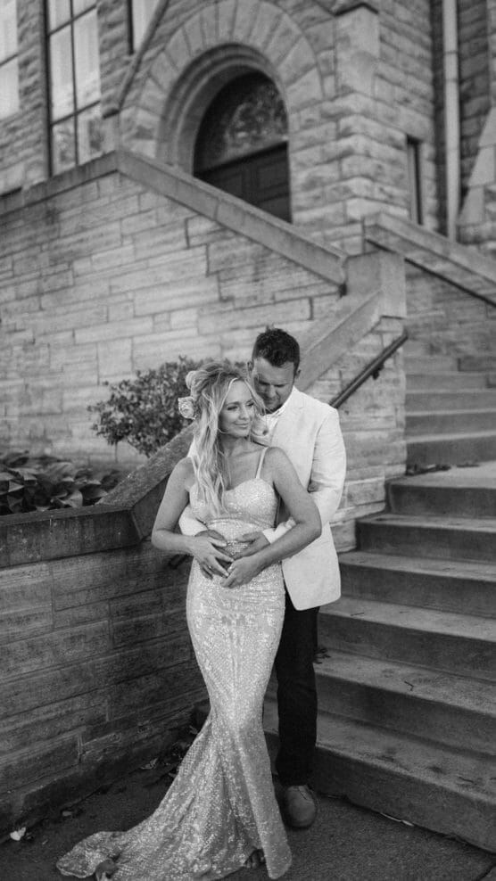 A couple poses in front of stone building steps. The woman wears a sparkling gown with her hair styled and flowers in it, while the man, in a light jacket, stands behind her, holding her around the waist. The image is in black and white. by Justin Salem Meyer