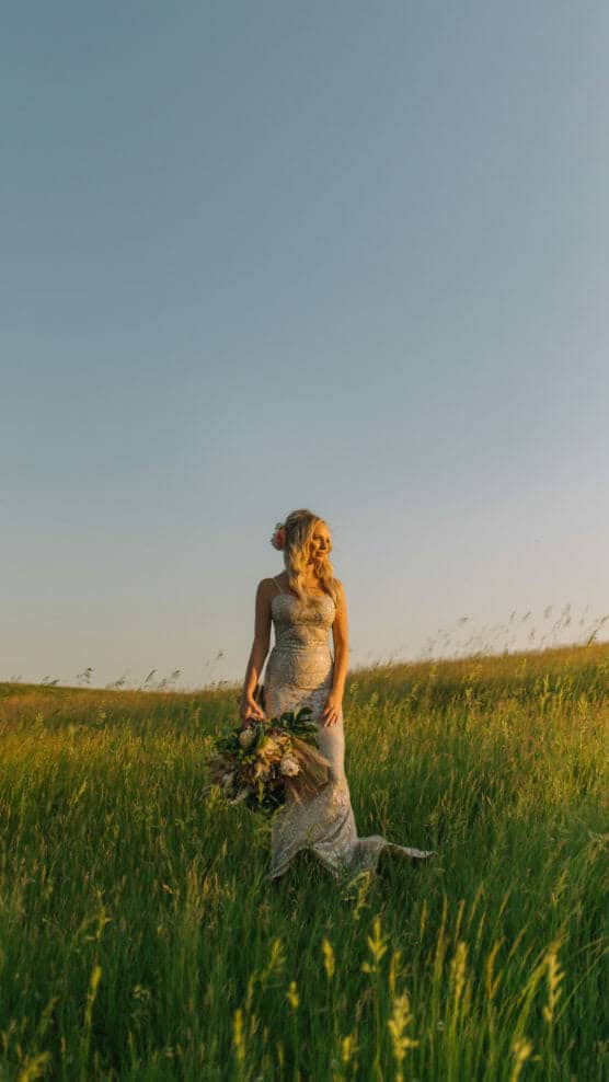 A woman in a sparkling dress stands in a sunlit grassy field, holding a bouquet of flowers, with clear blue sky and gentle sunlight creating a serene atmosphere. by Justin Salem Meyer