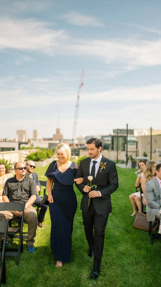 A man in a suit holding a white rose walks arm-in-arm with a woman in a navy dress down an outdoor aisle, surrounded by seated guests on a sunny day with city buildings and a crane in the background. by Justin Salem Meyer