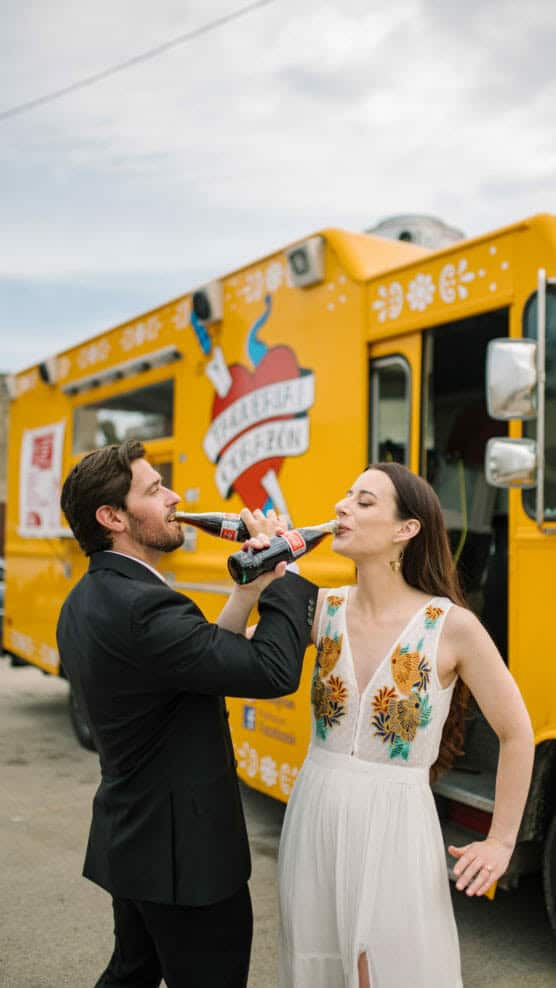 A man in a suit and a woman in a white dress with floral embroidery drink soda and laugh in front of a bright yellow food truck on a sunny day. by Justin Salem Meyer
