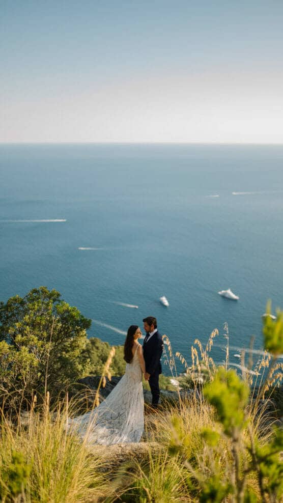 A bride and groom stand on a grassy hill overlooking the ocean, holding hands and gazing at each other, with boats visible in the blue water below under a clear sky. by Justin Salem Meyer