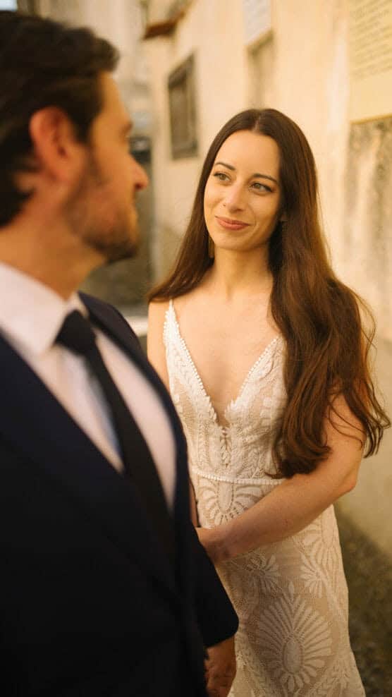 A bride in a white lace dress with long brown hair smiles warmly at a groom in a dark suit and tie as they stand together outdoors, holding hands. by Justin Salem Meyer