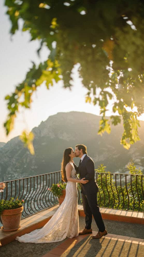 A couple in wedding attire shares a kiss on a scenic terrace at sunset, with mountains and a sunlit sky in the background and leafy branches framing the top of the image. by Justin Salem Meyer