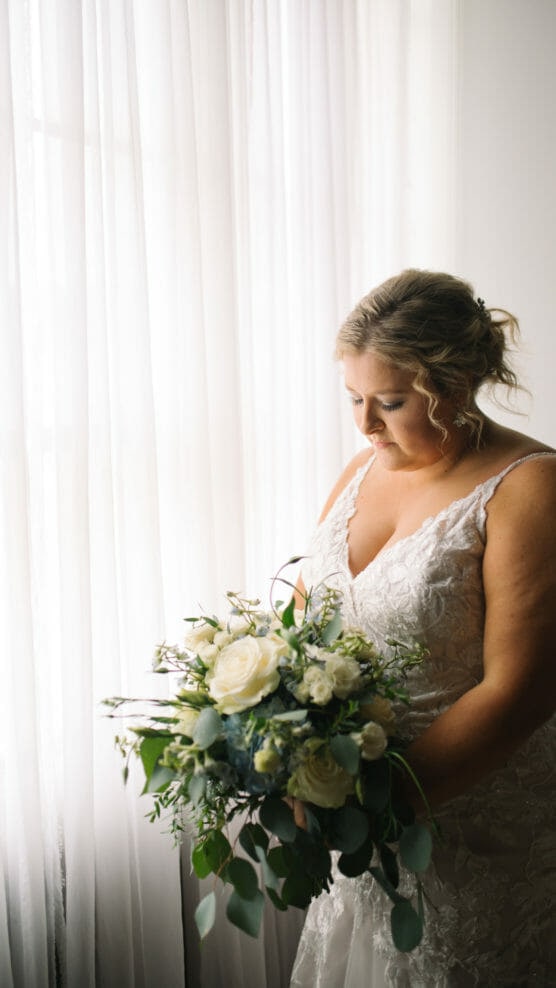 A bride in a white lace wedding dress stands by a window with sheer curtains, holding a bouquet of white roses and greenery, looking down thoughtfully. by Justin Salem Meyer