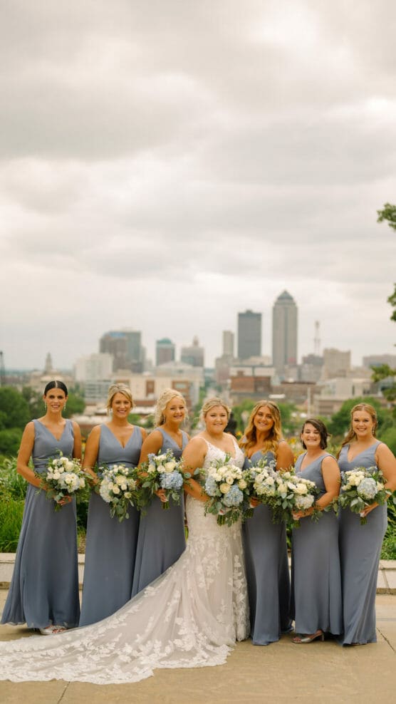 A bride in a white lace gown stands with six bridesmaids in matching slate blue dresses, each holding bouquets of white and green flowers, with a city skyline in the background under a cloudy sky. by Justin Salem Meyer