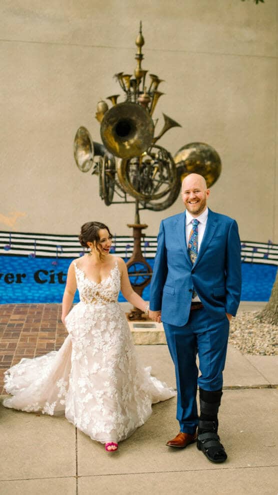 A bride in a white floral dress and a groom in a blue suit with a leg brace hold hands and smile while walking outdoors. A large sculpture made of brass instruments is in the background. by Justin Salem Meyer
