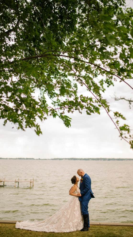 A bride and groom stand closely together by a lake under a large tree, with the bride in a long, flowing gown and the groom in a blue suit on a cloudy day. by Justin Salem Meyer