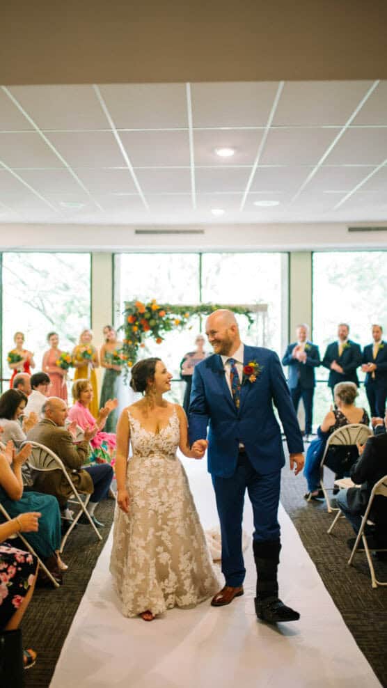 A bride in a white floral dress and a groom in a blue suit walk hand-in-hand down the aisle, smiling at each other, with guests seated on both sides and a floral arch in the background. by Justin Salem Meyer