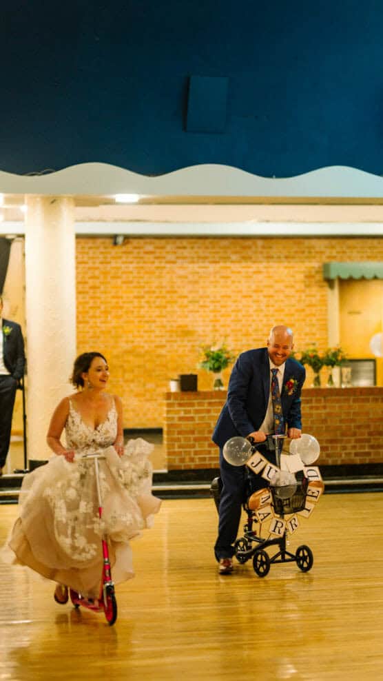 A bride in a white dress and a groom in a blue suit race on scooters indoors, both smiling and having fun on a polished wooden floor. The groom’s scooter is decorated with balloons and cans. by Justin Salem Meyer