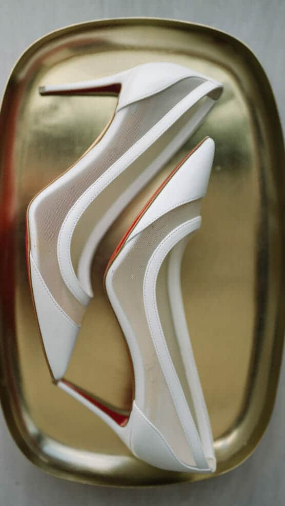 A pair of elegant white high-heeled shoes with transparent mesh panels, displayed side by side on a shiny gold tray. by Justin Salem Meyer