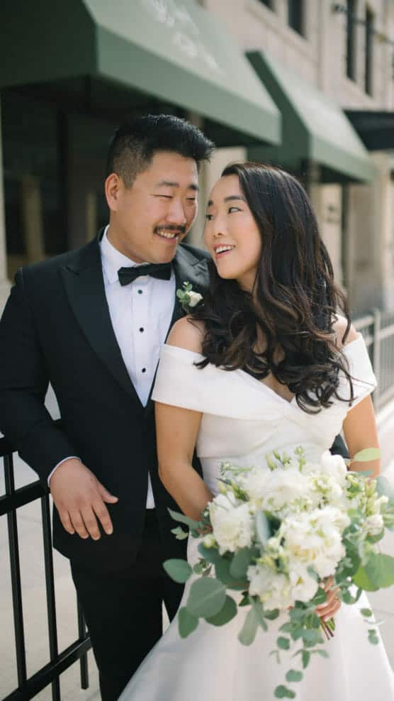 A groom in a black tuxedo smiles at his bride, who wears an off-the-shoulder white wedding dress and holds a bouquet of white and green flowers. They stand outdoors in front of a building with green awnings. by Justin Salem Meyer