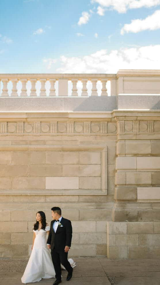 A bride in a white gown and a groom in a black tuxedo walk hand in hand along a light stone building under a blue sky with scattered clouds. by Justin Salem Meyer