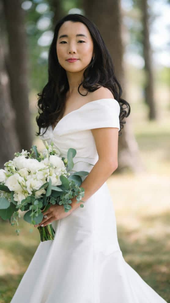 A bride with long dark hair wearing an off-the-shoulder white wedding dress holds a bouquet of white flowers and greenery, standing outdoors among trees on a sunny day. by Justin Salem Meyer