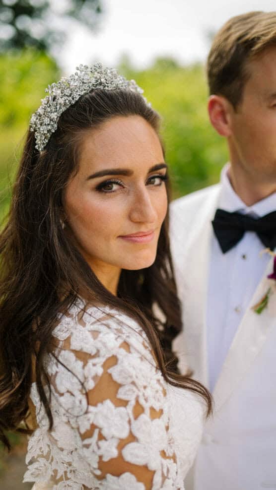 A bride with long brown hair, wearing a jeweled headpiece and a lace wedding dress, stands next to a groom in a white tuxedo jacket and black bow tie. The grooms face is partially out of frame. by Justin Salem Meyer