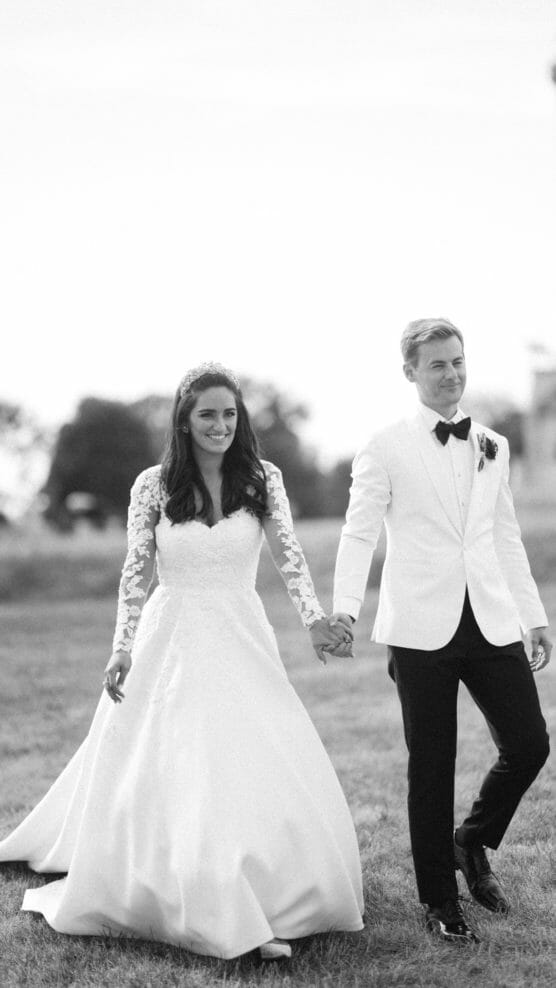 A bride in a long-sleeved lace wedding dress and a groom in a white tuxedo jacket and black bow tie walk hand in hand outdoors, smiling. by Justin Salem Meyer