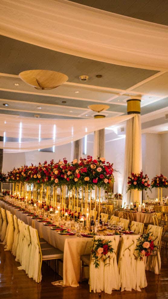 A lavishly decorated banquet hall features long tables with gold chairs, elegant white linens, tall floral centerpieces with red and pink flowers, and warm candlelight, creating a romantic, festive atmosphere. by Justin Salem Meyer