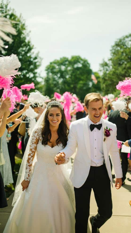 A smiling bride in a white lace wedding dress and veil walks hand-in-hand with the groom in a white tuxedo jacket. They are surrounded by guests waving pink and white pom-poms outdoors on a sunny day. by Justin Salem Meyer