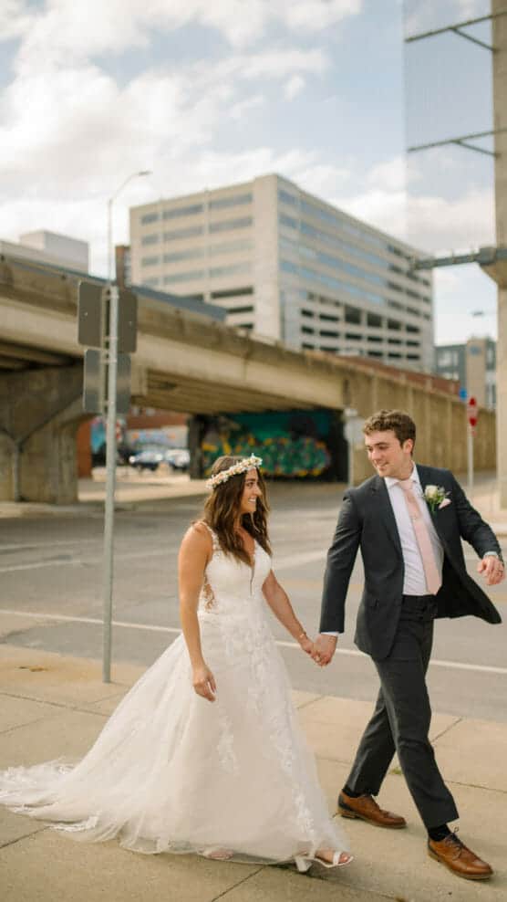A bride in a white gown and floral crown holds hands with a groom in a suit as they walk outside on a city sidewalk, smiling at each other with buildings and an overpass in the background. by Justin Salem Meyer