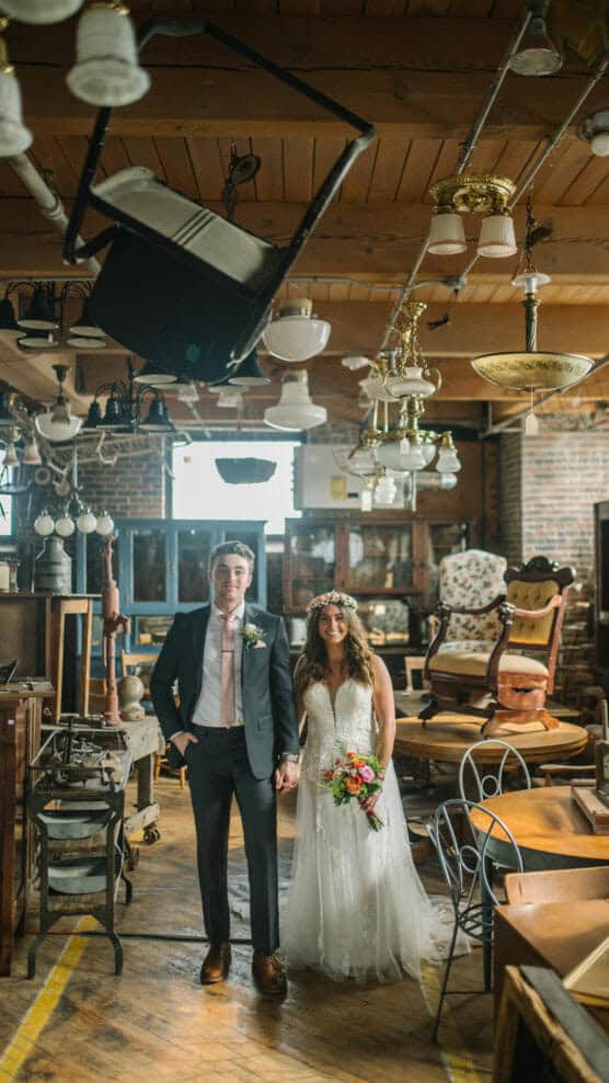 A bride and groom stand holding hands in a vintage shop filled with antique furniture and light fixtures, smiling at the camera. The bride wears a floral crown and white dress; the groom is in a suit with a pink tie. by Justin Salem Meyer