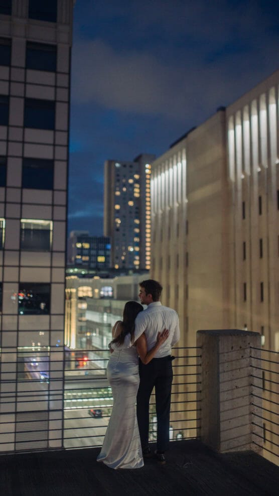 A couple stands close together on a balcony at night, facing away and looking at a cityscape with tall, illuminated buildings under a dark, cloudy sky. The woman wears a white dress; the man wears dark pants and a white shirt. by Justin Salem Meyer