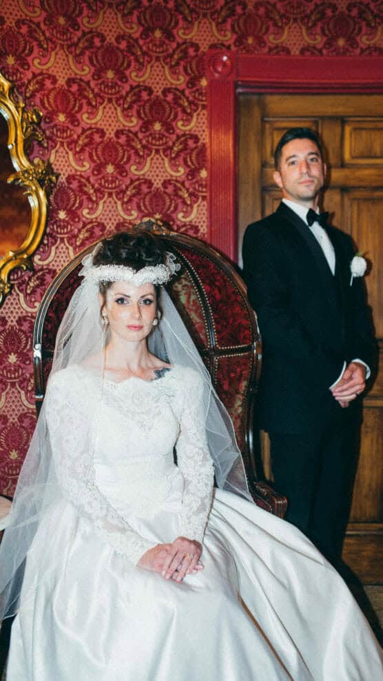 A bride in a white gown and veil sits on an ornate chair, while a groom in a black tuxedo stands beside her. The room has patterned red wallpaper, a wooden door, and a decorative mirror. by Justin Salem Meyer