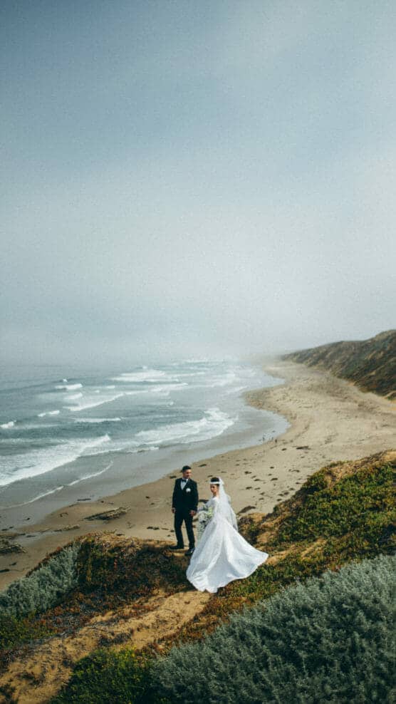 A bride and groom stand together on a grassy cliff overlooking a deserted beach and ocean waves under a cloudy sky, with the bride’s white dress flowing in the breeze. by Justin Salem Meyer