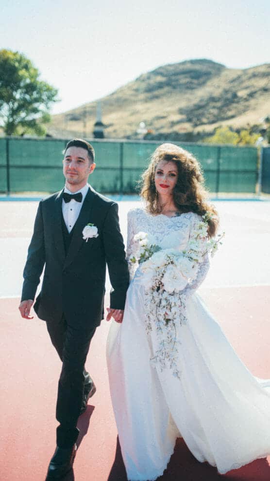 A bride in a white wedding dress holding a bouquet walks hand in hand with a groom in a black tuxedo on an outdoor court, with hills and trees in the background on a sunny day. by Justin Salem Meyer