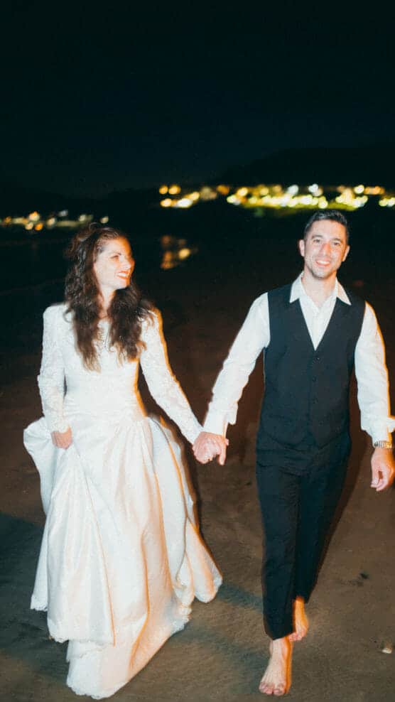 A smiling couple, dressed in formal attire, walks barefoot on a beach at night, holding hands. The woman wears a long white dress, and the man wears a white shirt with a dark vest and pants. City lights glow in the background. by Justin Salem Meyer