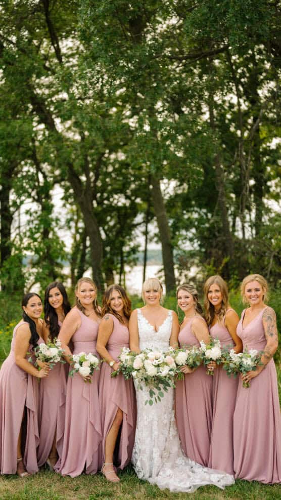 A bride in a white lace gown stands outdoors with seven bridesmaids in matching mauve dresses, all holding white and green bouquets, smiling in front of tall green trees. by Justin Salem Meyer